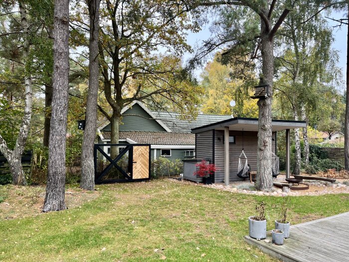 Trädgård med vedbod, hammockar och stenlagd eldplats under tallar. Pepparmyntsgröna hus i bakgrunden bland höstfärgade träd.