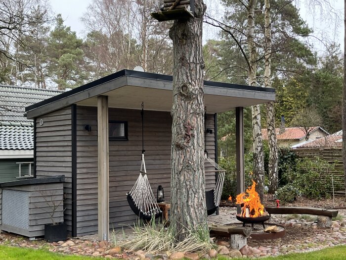 Uteplats med vedförråd, hängmatta och en liten eld i en eldkorg. Vedträ bredvid, omgiven av grönska och träd.