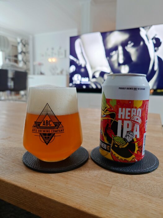 Glas med Apex Brewing Company-logo fyllt med IPA-bärnstensöl bredvid en burk med etikett "Hero IPA" på träbord.