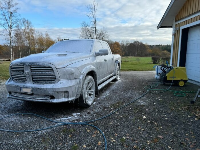 Bil täckt med rengöringsskum på uppfart bredvid garage och tvättutrustning, omgiven av höstträd.