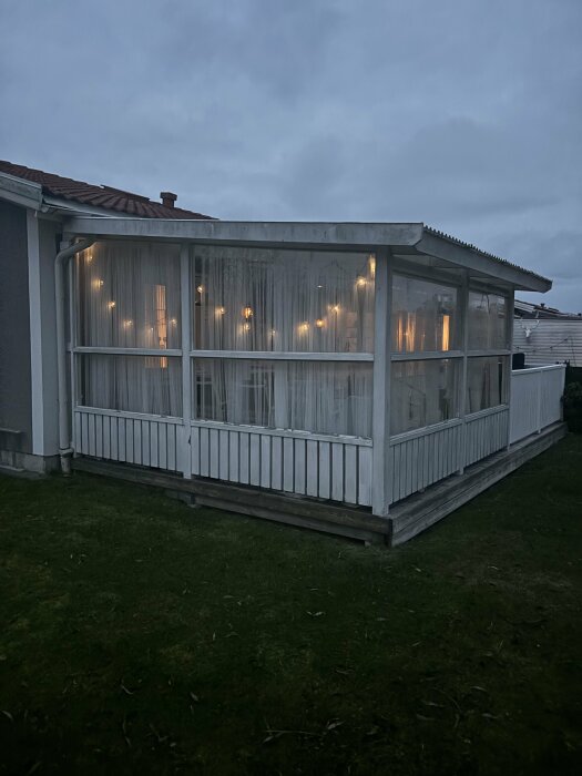 Altan med transparanta gardiner och belysning, ansluten till hus med rött tak. Stuprör syns vid sidan av altanen, belägen på gräsmatta under mulen himmel.