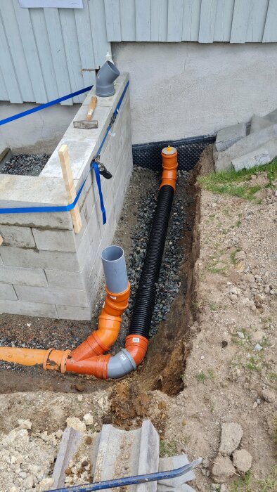 Dräneringsarbete vid grundmur med synliga avloppsrör, grå och orange, som leder bort vatten. Grus täcker rören och en mur av betongblock syns i bakgrunden.