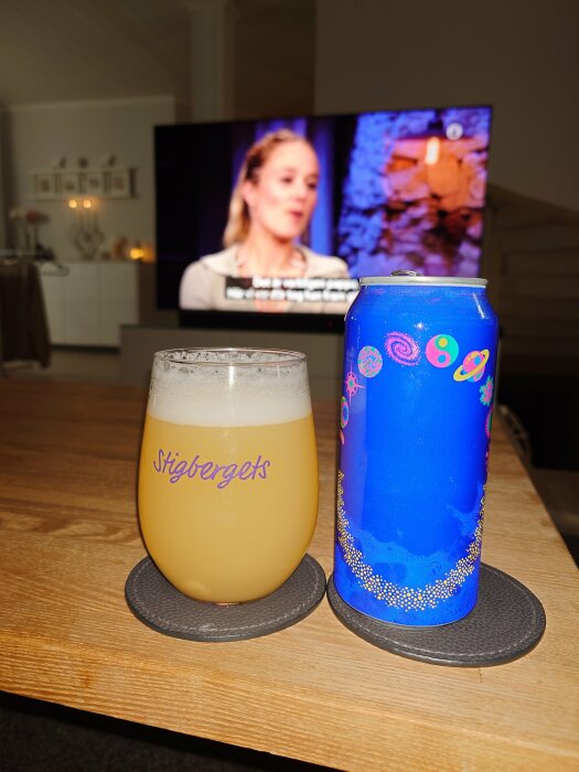 En öl i ett Stigbergets-glas och en blå ölburk på ett bord, med en påslagen TV i bakgrunden.