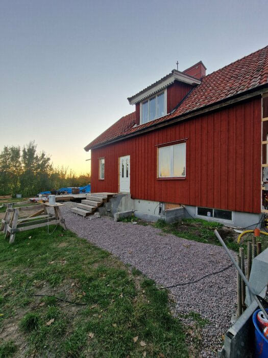 Rött hus med brunt tak, grusad gång leder till trappa. Saknar bräfoder och knutar, framtida projekt inkluderar altan och källaren.