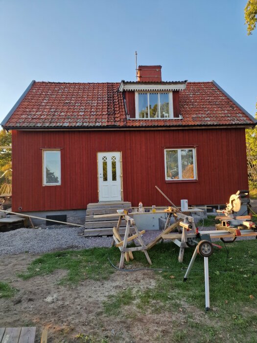 Rött hus under renovering med brädor och verktyg utanför, saknar brädfoder och knutar. Solig dag.