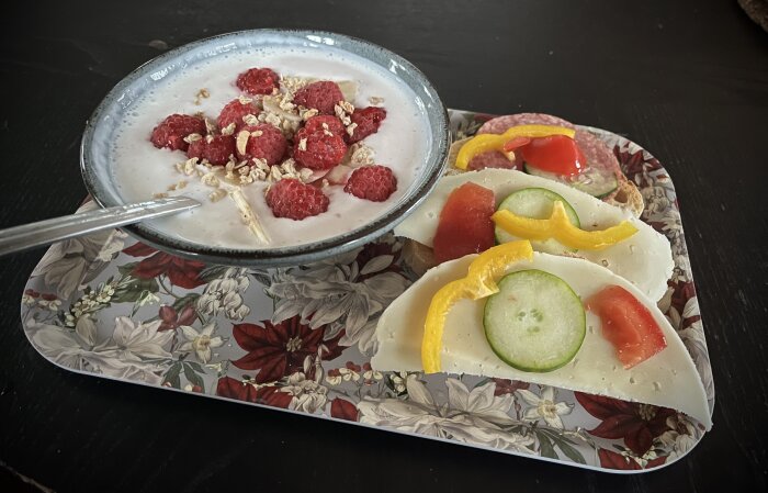 Frukost med en skål fil toppad med hallon och müsli, samt smörgåsar med ost, gurka, paprika och tomat på en blommig bricka.