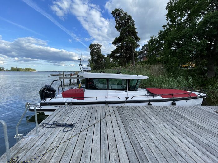 Vit motorbåt med röd täckning ligger förtöjd vid en brygga, omgiven av grönska och vatten under en blå himmel med moln.