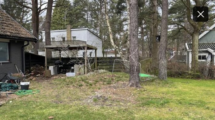 Trädgård med buskar, träd och en uteplats. En hög med grästoppar och växter under förändring, med hus i bakgrunden.