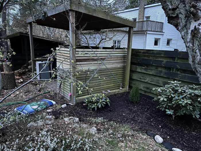 Trädgård med nyplanterad rhododendron, liten stengång och träpaviljong. Plantering omgiven av trästaket och betongkant.