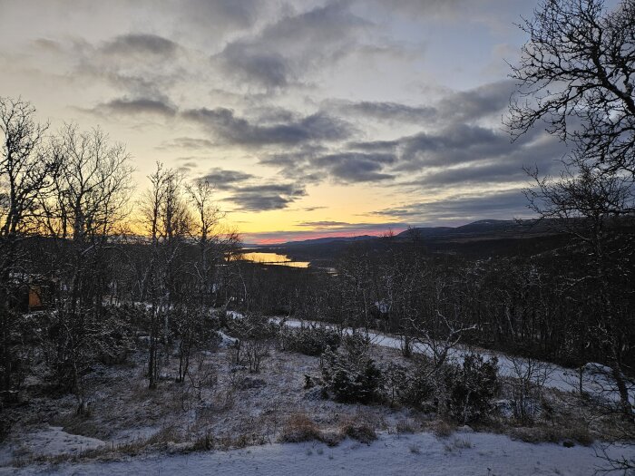 Solnedgång över snötäckt landskap med träd och berg, och en vägg av moln på himlen i bakgrunden.