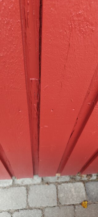 Närbild av rödmålad träfasad med sprickor och ojämn färgyta, belägen ovanför en stenlagd yta.