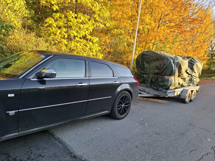 Svart bil med släpkärra lastad med täckt ved på en höstväg. Gulbladen träd i bakgrunden.