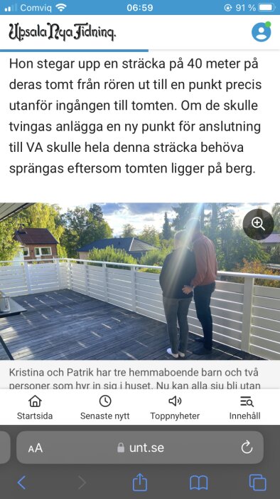 Två personer står på en altan med vita staket och en trädgård i bakgrunden under en solig himmel.