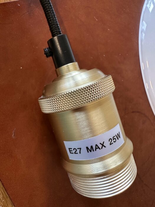 E27 lampsockel med metallfinish och text "E27 MAX 25W", ansluten till en svart kabel, visas på en brun yta.