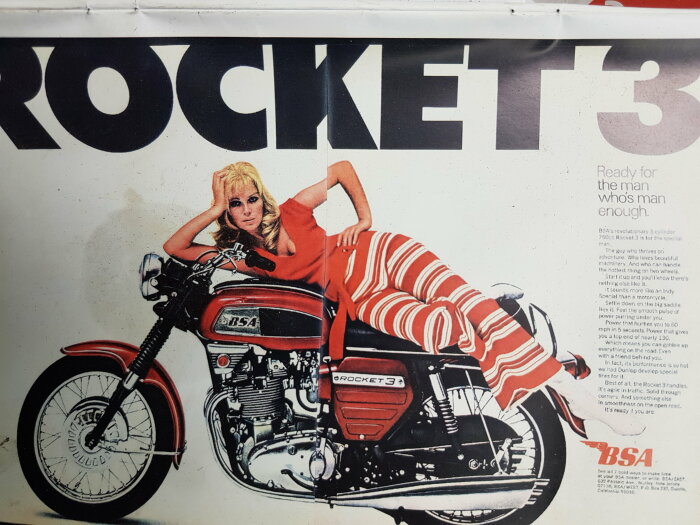 Reklamaffisch med en kvinna i randig klädsel liggande på en röd BSA Rocket 3-motorcykel, text och logotyp synliga.