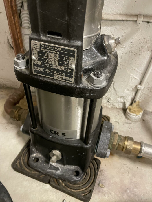 Grundfos CR5 vattenpump i källare, med etikett som visar specifikationer och anslutna rörledningar. Pumpen orsakar kontinuerligt varmvattenproblem.