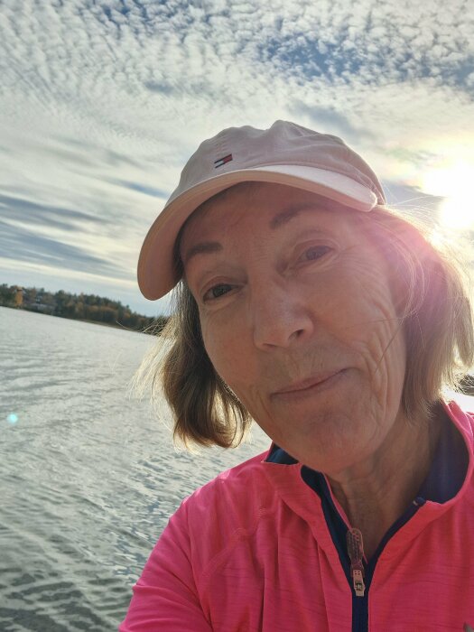 Person i rosa jacka och mössa tar en selfie vid vatten, en solig dag med molnig himmel i bakgrunden.