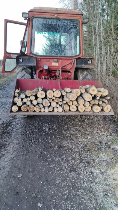 En traktor med en skopa full av småved vid en grusväg i skogen.