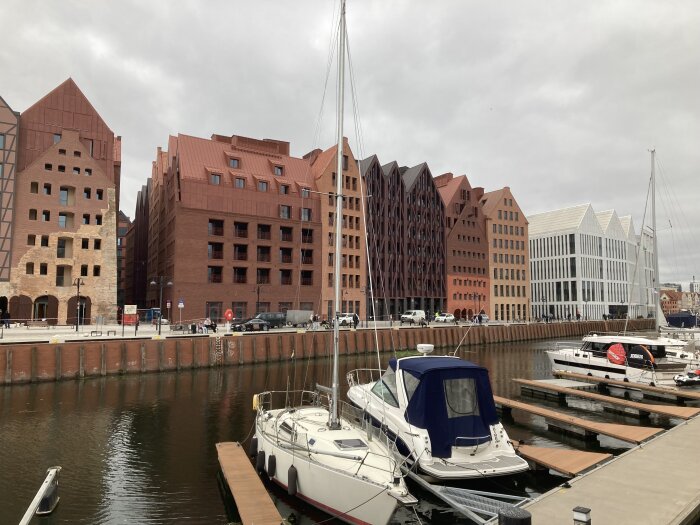 Modern byggnadskomplex vid vattnet med båtar förtöjda vid bryggor i förgrunden och molnig himmel i bakgrunden.