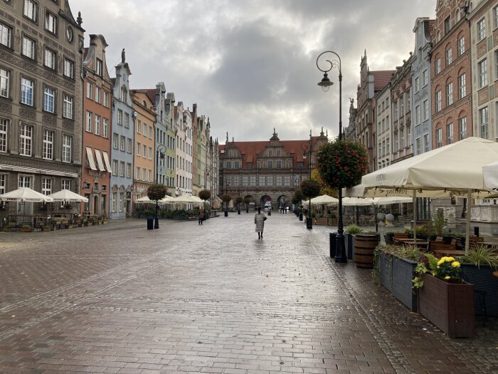 Gata kantad med äldre byggnader och uteserveringar under molnig himmel, med människor promenerande i bakgrunden.