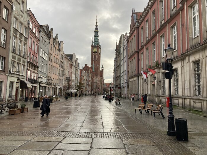 Gata med färgglada, historiska byggnader och ett klocktorn på en mulen dag. Människor promenerar längs trottoaren.