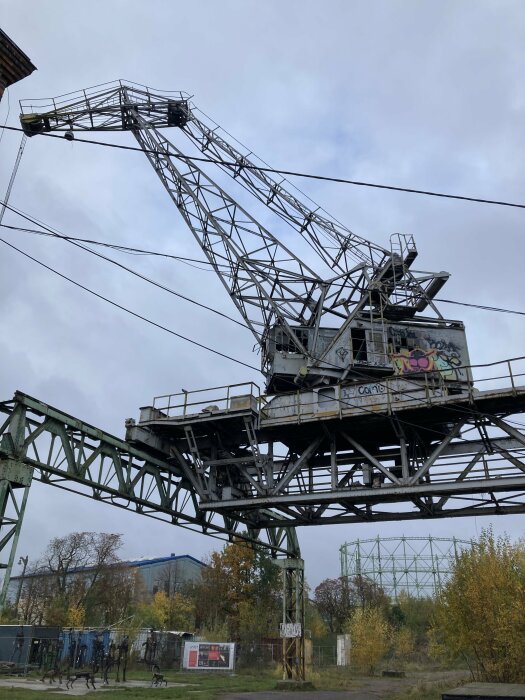 Äldre industriell kran med metallstruktur och graffitimålningar, placerad i ett övergivet område med träd och industribyggnader i bakgrunden.