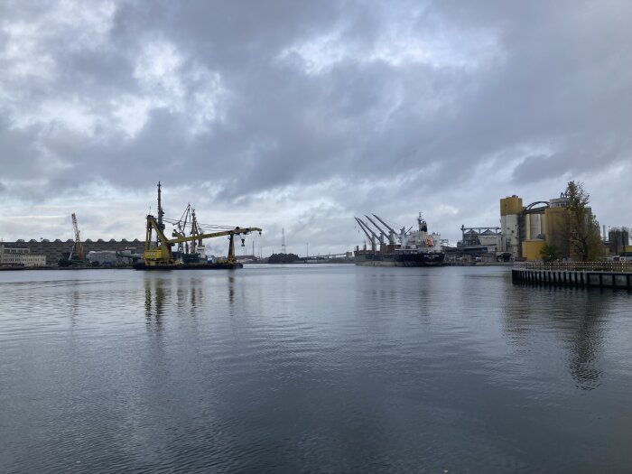 Industriell hamn med lastkranar och fartyg vid disigt väder, stilla vatten i förgrunden och fabriksbyggnader i bakgrunden.