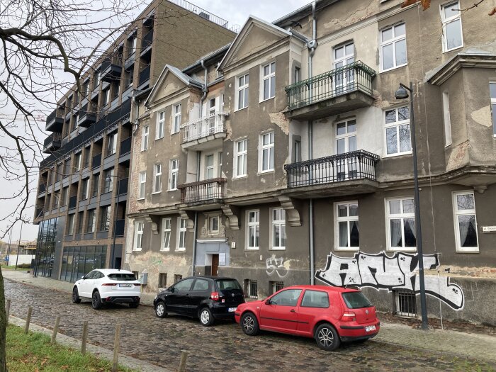 Gatuliv framför äldre bostadshus med graffiti på fasaden, tre bilar parkerade längs stenlagd gata, vintertid.