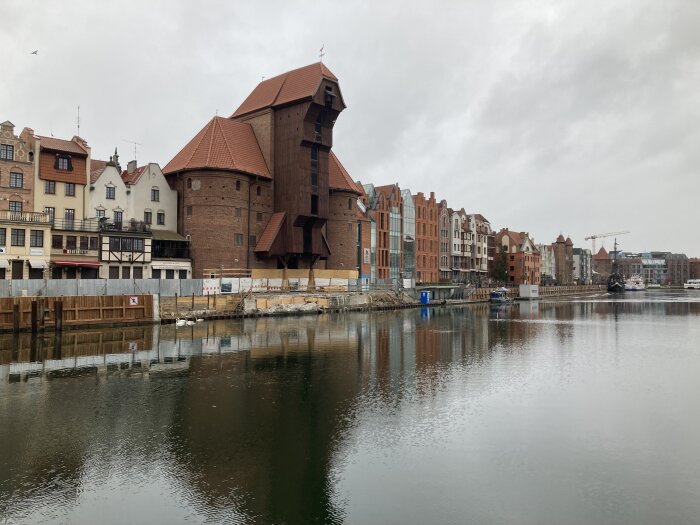 Historiska byggnader vid en flod med reflektioner i vattnet, en gammal stadsmiljö med tegelarkitektur och trästrukturer under mulen himmel.