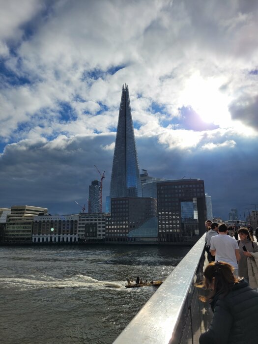 The Shard i London under en molnig himmel, med människor som promenerar på en bro i förgrunden och en båt som färdas på floden nedanför.