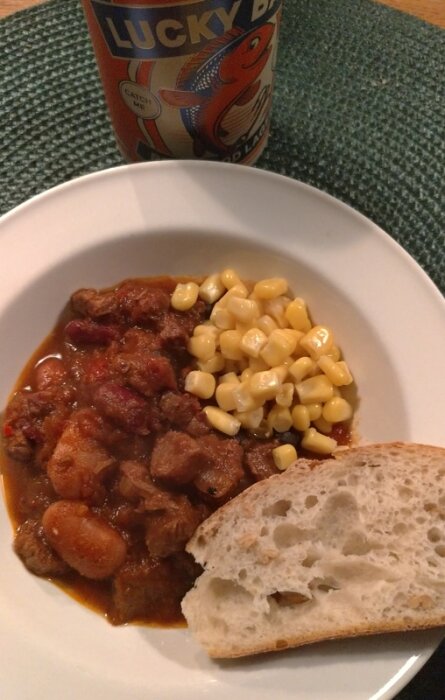 En skål med hemlagad chili med bönor och majs, serverad med bröd, och en läskburk i bakgrunden.