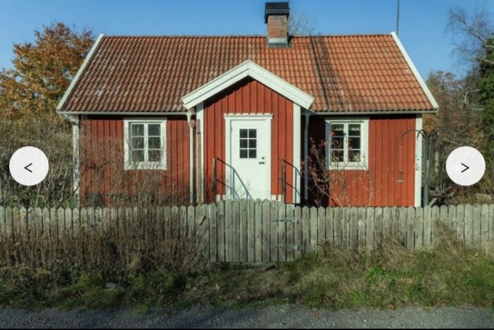 Rött trähus med tegeltak, liten förstukvist och två fönster, omgivet av trästaket och höstfärgade buskar, fotograferat framifrån.