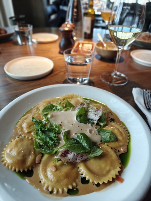 Hummerravioli på en vit tallrik med gröna blad och skum, serverad på en träbord med dryck i bakgrunden.