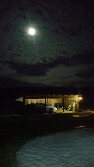 Nattbild av upplyst carport med bil under, fullmånen lyser bland moln, snötäckt mark i förgrunden.