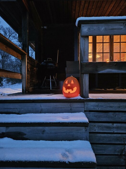 Upplyst Jack-o'-lantern på snötäckt veranda med stearinljus och utemöbler i bakgrunden, under skymmning.