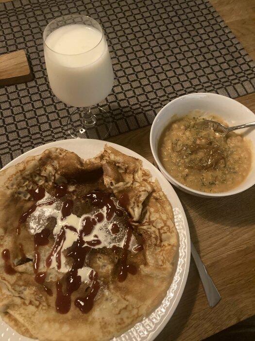 Pannkakor med sylt och grädde på en tallrik, ärtsoppa i en skål och ett glas mjölk på ett bord med grå duk.