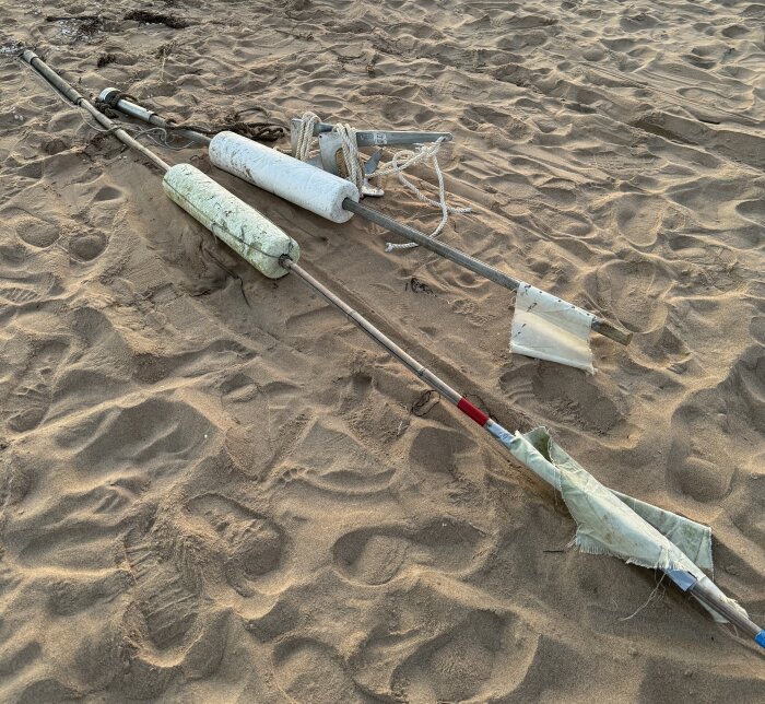 Två fiskenätvakare med flytelement ligger på en sandstrand, med rep och plastöverdrag synliga.