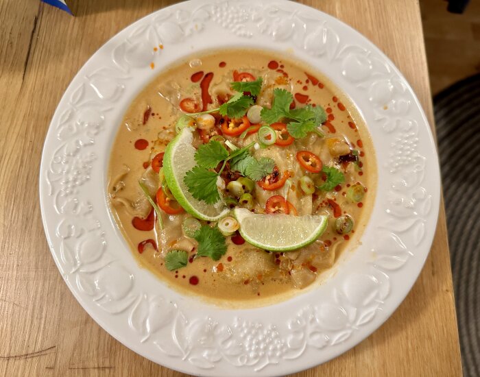 Dumplingsoppa med chili, koriander, salladslök, lime och chiliolja i en vit skål på ett träbord.