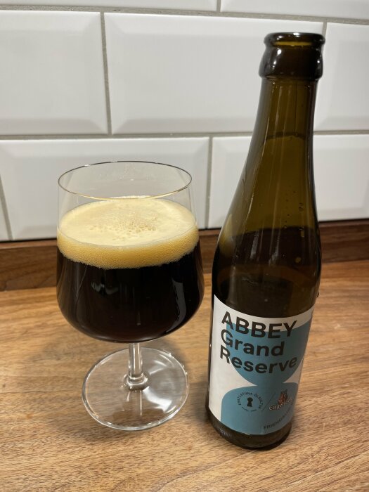 Flaska och glas med mörk öl märkt Abbey Grand Reserve står på en träbänk framför vita kakelplattor.