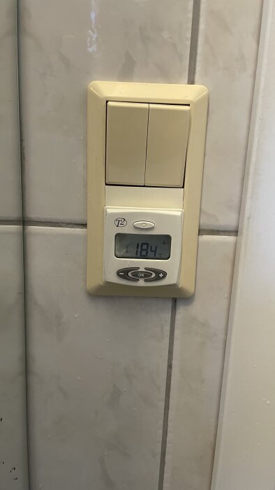 Gulnad väggbrytare med vit termostat på kakelvägg, visar 18,4°C.