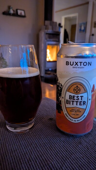 Glas med mörk öl och ölburk märkt "Buxton Brewery Best Bitter" på bord, brasa i bakgrunden.