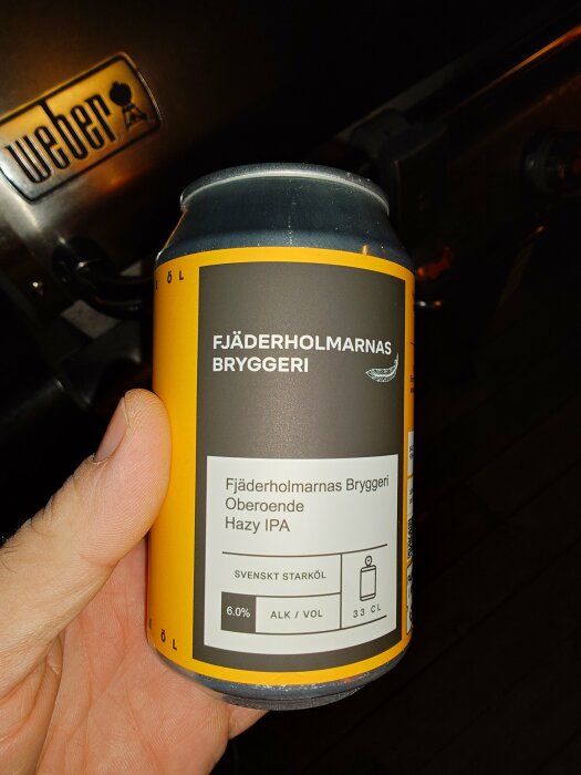 En hand håller en ölburk från Fjäderholmarnas Bryggeri med etiketten "Oberoende Hazy IPA" framför en Weber-grill.