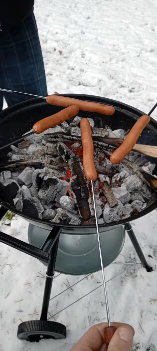 Korvgrillning på snötäckt mark över kolgrill. Flera korvar träs på grillspett och grillas över glödande kol.