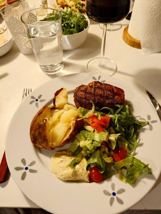 En tallrik med bakad potatis, grillad oxfilé, sallad och bearnaissås, omgiven av ett glas vin och vatten på ett dukat bord.