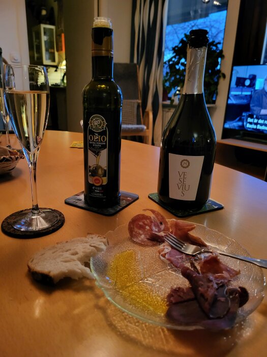 Charkbricka med olivolja, bröd, salami och parmaskinka på tallrik. Proseccoglas och olivoljeflaska i bakgrunden på ett träbord.