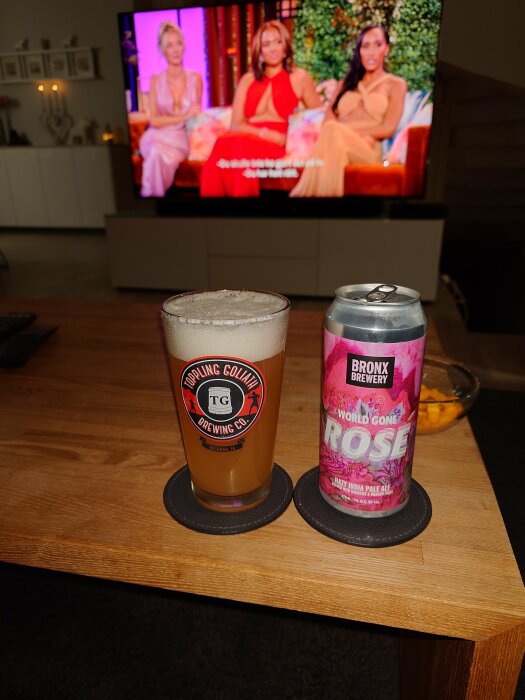 En ölburk från Bronx Brewery och ett glas öl på ett bord framför en TV som visar ett program med tre kvinnor.