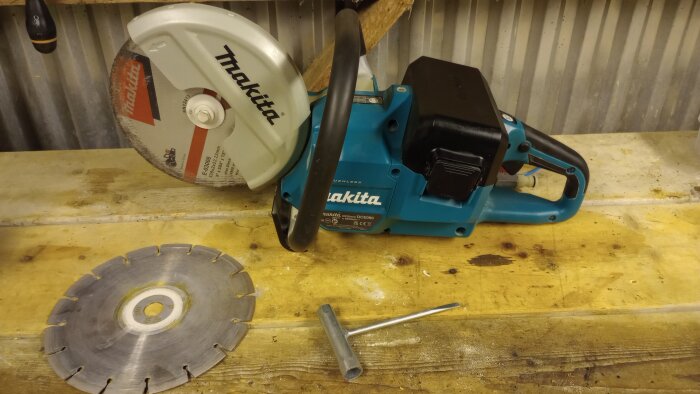 Makita batteridriven kapmaskin med kapskiva på 230mm och handtag, liggandes på en träbänk, diamantskiva och verktyg bredvid.