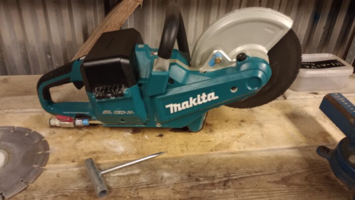 Makita batteridriven kapmaskin med 230mm skiva, används för kapningar i betong, asfalt och metall. Verktyg och kapskiva syns intill på arbetsbänk.