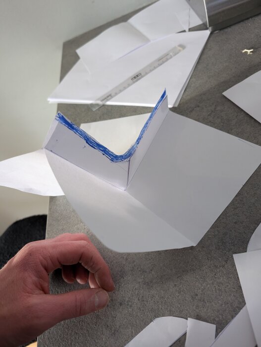 Paper model of an angled structure on a table with a hand holding it, surrounded by scattered papers and a ruler.