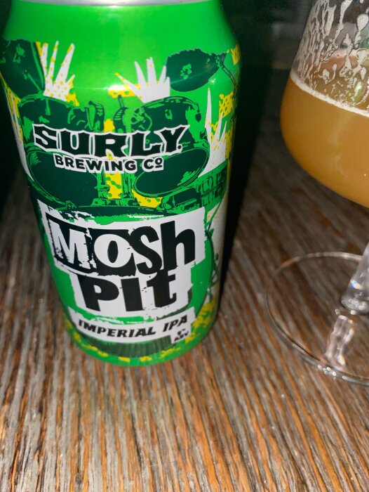 Grön burk av Mosh Pit Imperial IPA från Surly Brewing, bredvid ett halvfullt glas med öl, placerad på ett träbord.